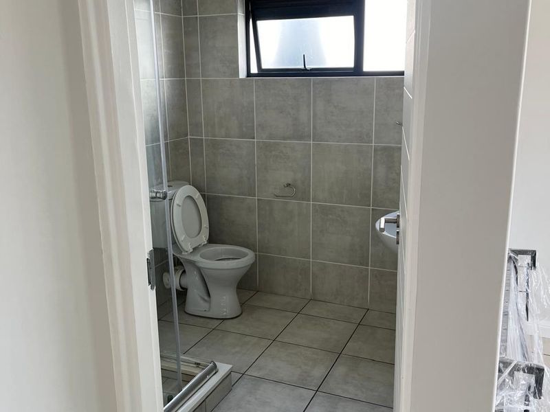 A clean, modern bathroom featuring neutral grey floor and wall tiles, a pedestal basin, a toilet, and a glass-enclosed shower.