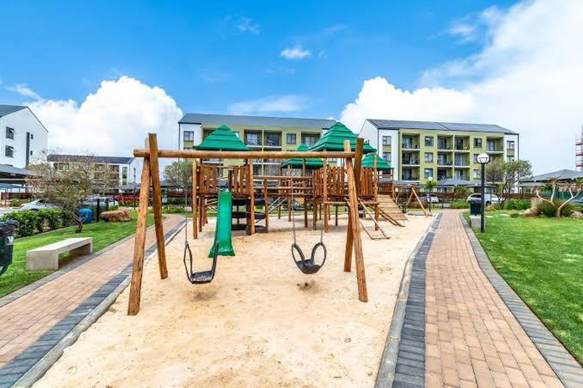 A well-maintained communal playground area featuring wooden play structures, swings, and paved pathways set within a landscaped residential apartment complex garden.