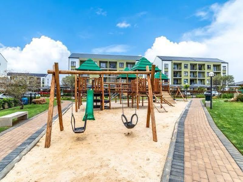A well-maintained communal playground area featuring wooden play structures, swings, and paved pathways set within a landscaped residential apartment complex garden.