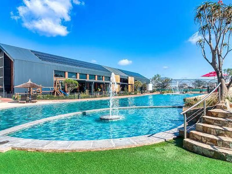 A well-maintained communal swimming pool area featuring a central water fountain, stone steps with stainless steel railings, and a modern building with solar panels.