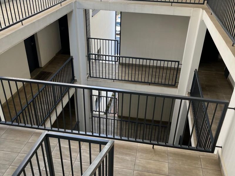 An open-air apartment complex hallway featuring tiled walkways, metal safety railings, and a central stairwell providing access to multiple units.