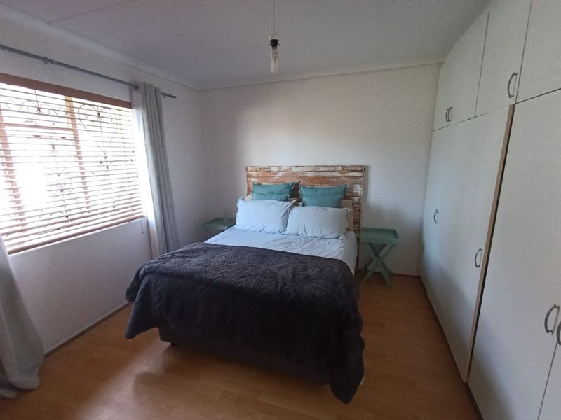 A functional bedroom featuring laminate flooring, built-in cupboards, and a window fitted with venetian blinds and security bars.