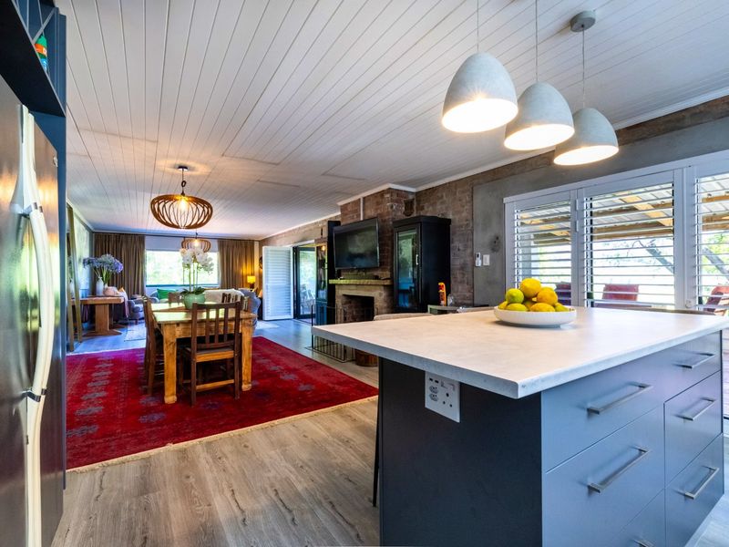 Spacious open-plan living and dining area featuring a modern kitchen island, brick fireplace, and timber-paneled ceilings with integrated pendant lighting.