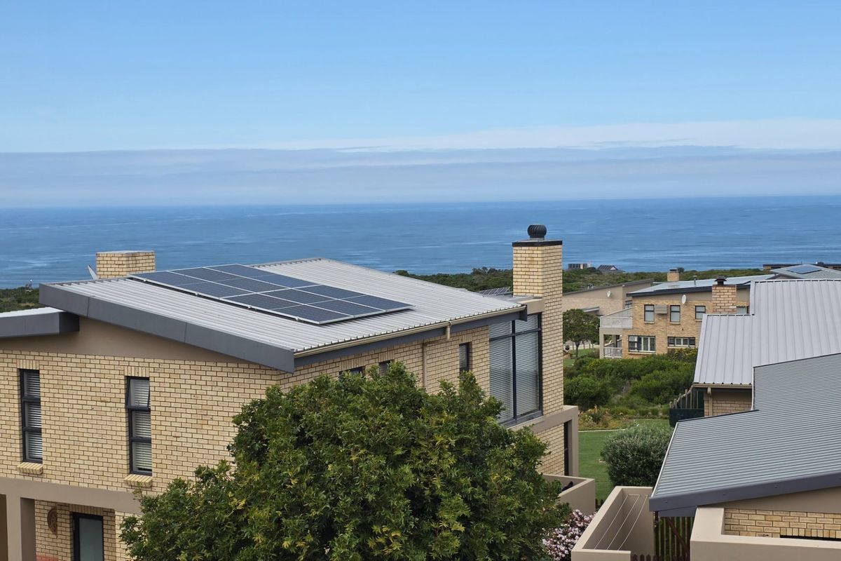 An elevated view of a modern brick house featuring a metal roof with integrated solar panels and a scenic coastal backdrop.
