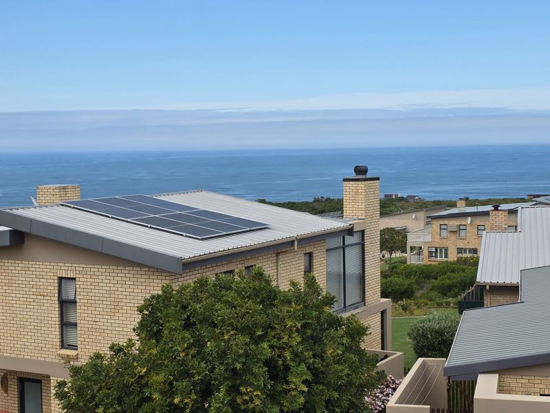 An elevated view of a modern brick house featuring a metal roof with integrated solar panels and a scenic coastal backdrop.