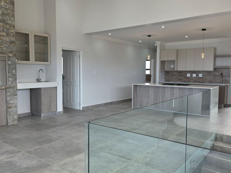 Spacious open-plan living area featuring a modern kitchen with island, built-in braai with stone cladding, and contemporary glass balustrade staircase.