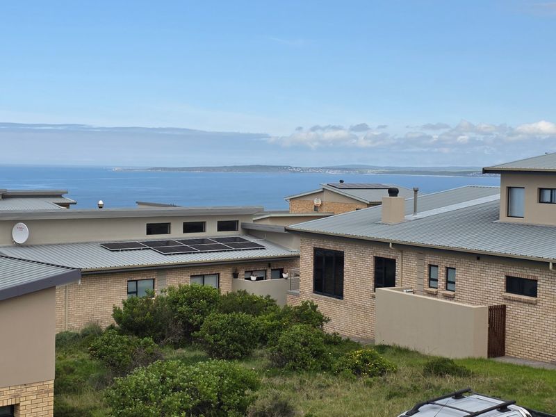 Elevated view overlooking a residential estate with modern brick homes, metal roofing, and solar panels, featuring a scenic coastal backdrop.