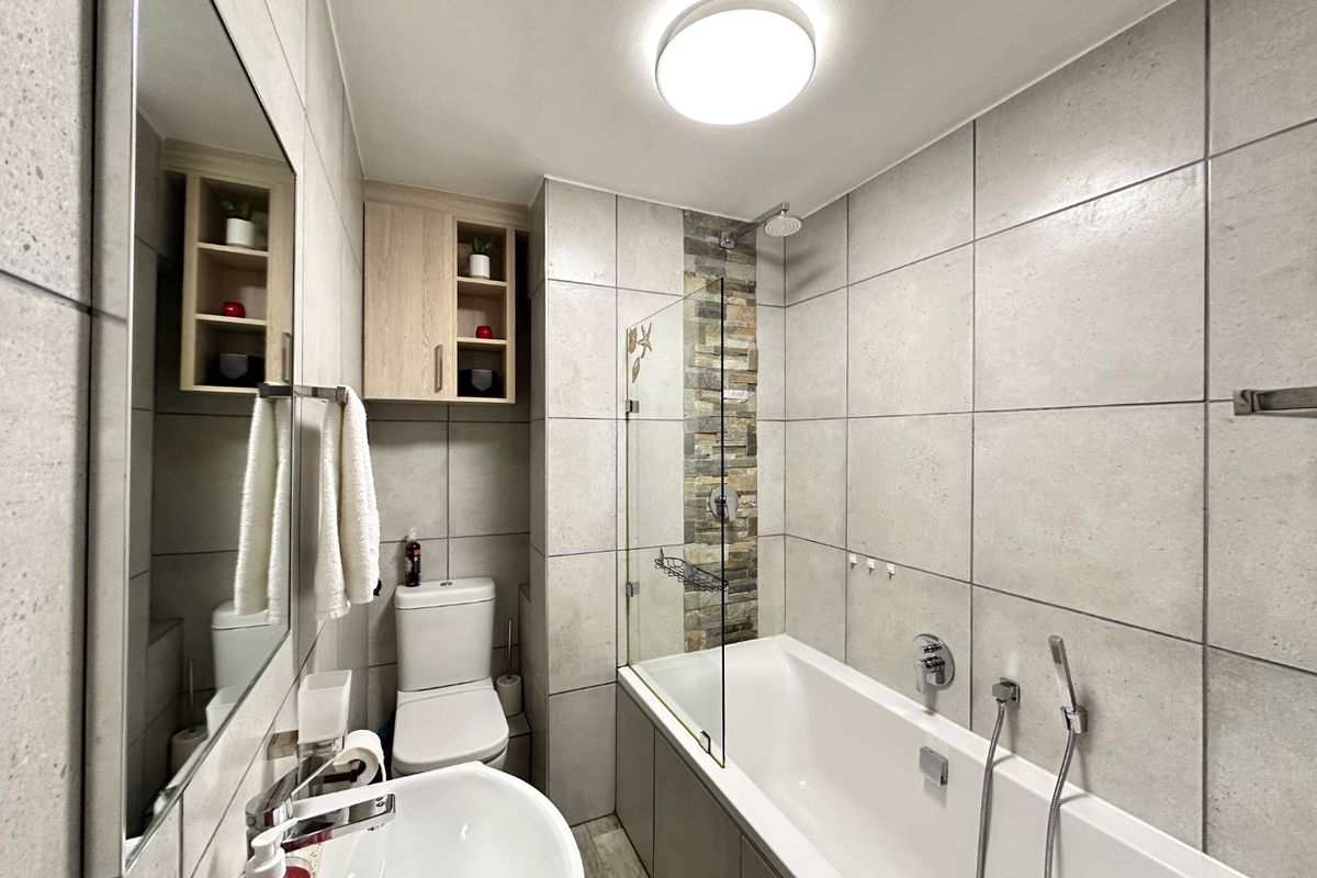 Modern bathroom featuring a bathtub with a glass shower screen, stone-cladding accent wall, built-in vanity storage, and neutral floor-to-ceiling tiling.