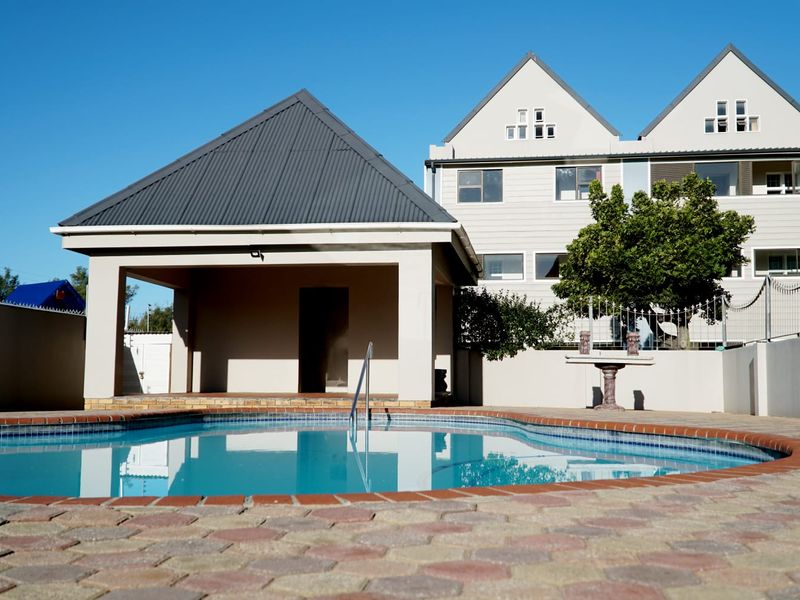 A well-maintained outdoor pool area featuring a paved deck, a covered patio structure, and secure boundary walls with electric fencing.