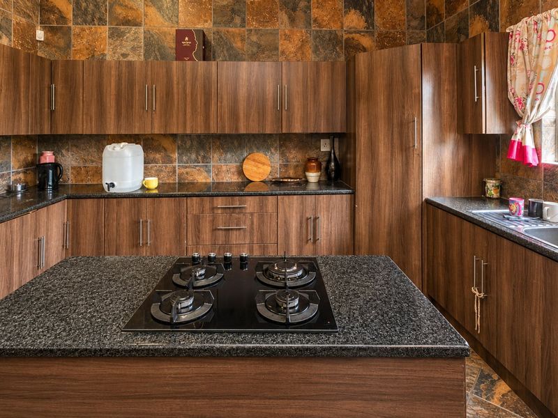 A functional kitchen featuring wood-finish cabinetry, dark granite countertops, a central island with a gas hob, and decorative stone-look wall tiling.