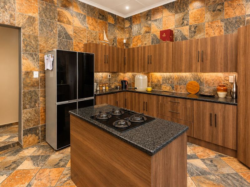 A well-maintained kitchen featuring wood-finish cabinetry, granite countertops, a central island with a gas hob, and distinctive stone-look wall and floor tiling.