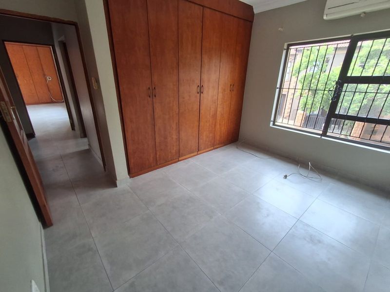 An empty bedroom featuring tiled flooring, built-in wooden cupboards, and a wall-mounted air conditioning unit, with security bars on the window.