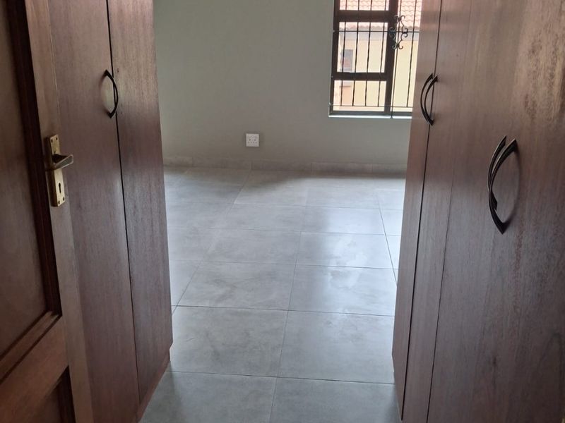 A clean, empty bedroom featuring neutral tiled flooring, built-in wooden cupboards, and a window fitted with security bars.