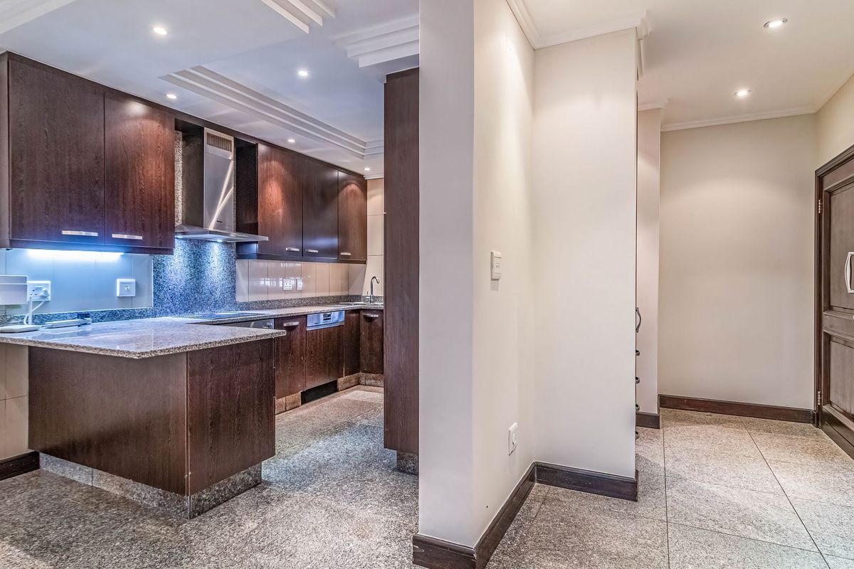 A modern kitchen featuring dark wood cabinetry, granite countertops, stainless steel extractor fan, and recessed lighting, with an adjacent tiled hallway area.