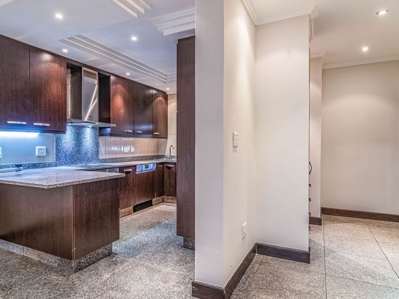 A modern kitchen featuring dark wood cabinetry, granite countertops, stainless steel extractor fan, and recessed lighting, with an adjacent tiled hallway area.