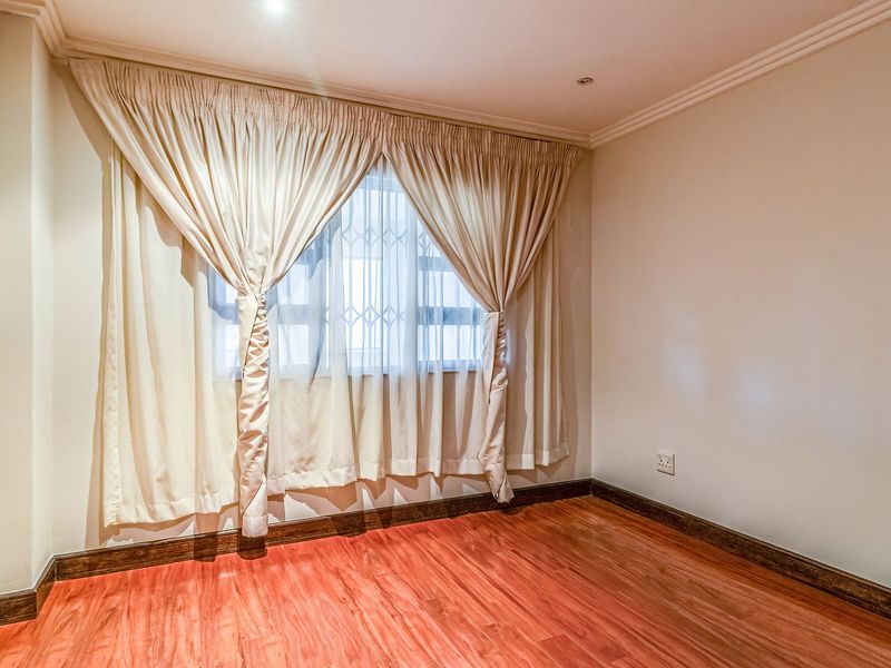 A bright, empty bedroom featuring warm-toned laminate flooring, recessed ceiling lighting, and a window fitted with a security gate and curtains.