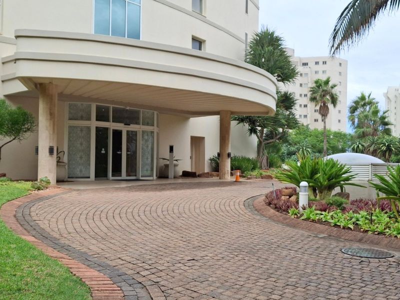 A well-maintained front entrance featuring a paved circular driveway, a covered portico with stone columns, and established garden landscaping.