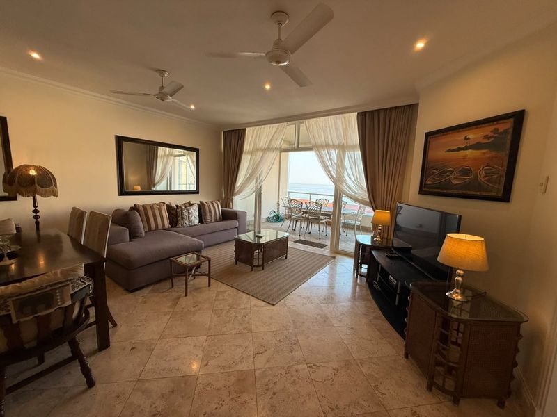 Spacious open-plan living and dining area featuring stone tile flooring, ceiling fans, and large sliding doors leading to a balcony with ocean views.