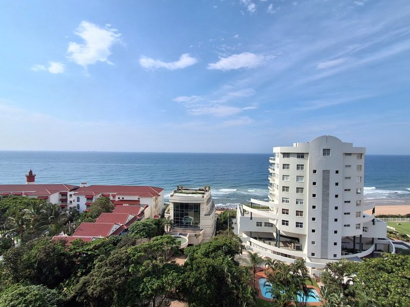 A scenic elevated view overlooking the coastline, featuring nearby residential buildings, lush greenery, and the ocean horizon.
