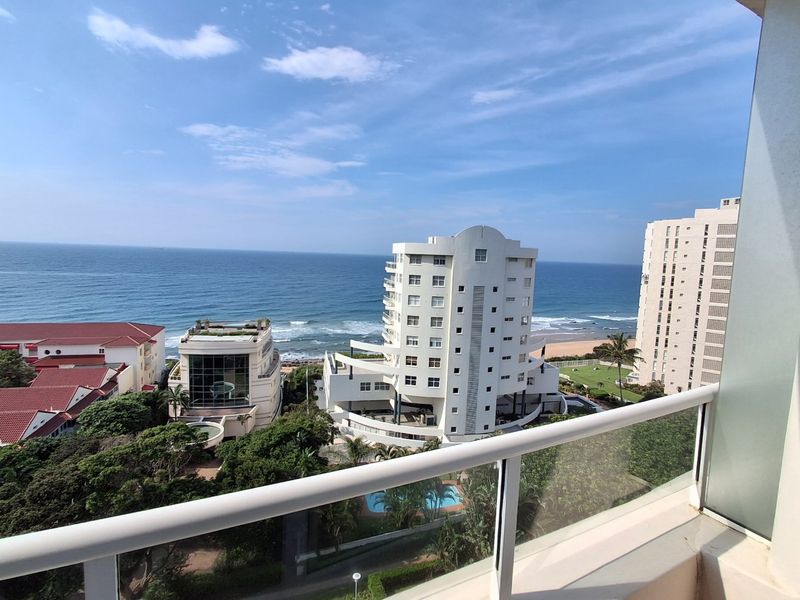 Elevated coastal view overlooking the ocean, surrounding apartment buildings, and landscaped grounds from a private balcony with a modern glass balustrade.