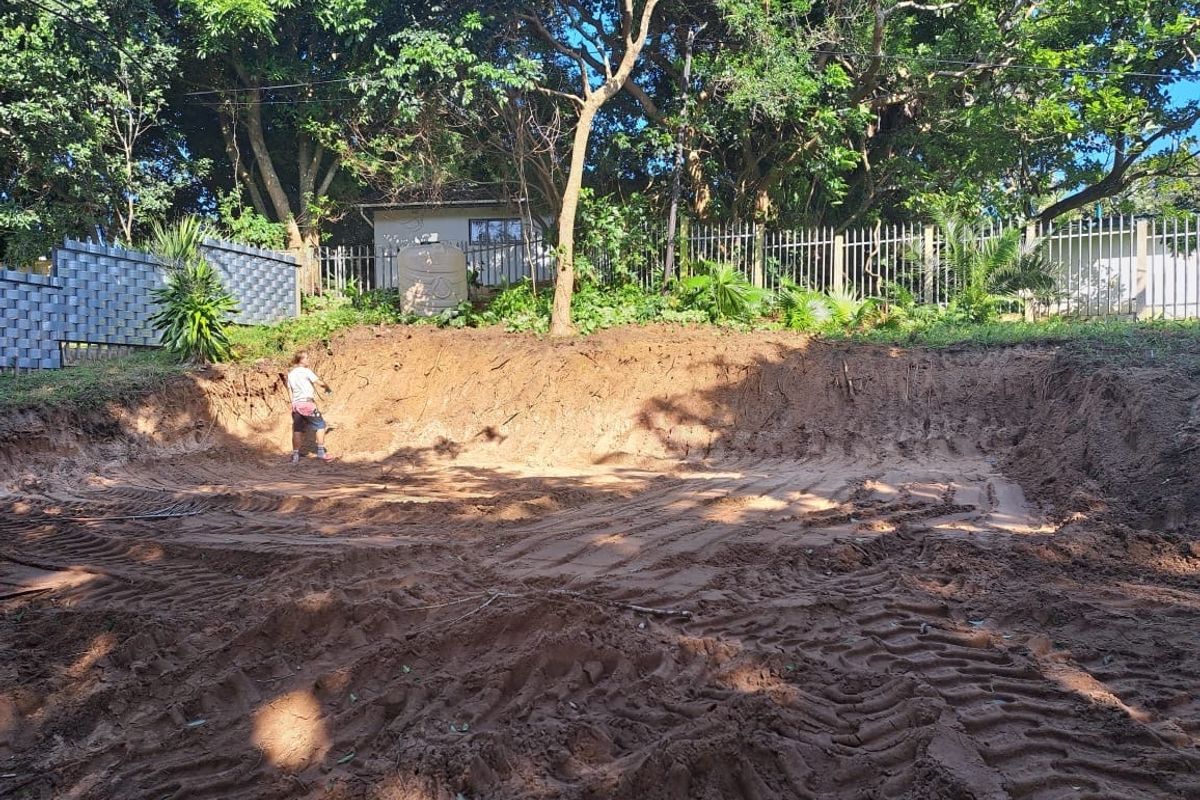 A vacant residential plot featuring significant earth excavation, mature trees, and perimeter metal fencing, suitable for future landscaping or development.