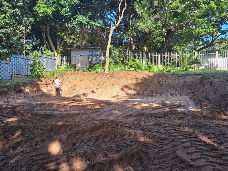 A vacant residential plot featuring significant earth excavation, mature trees, and perimeter metal fencing, suitable for future landscaping or development.
