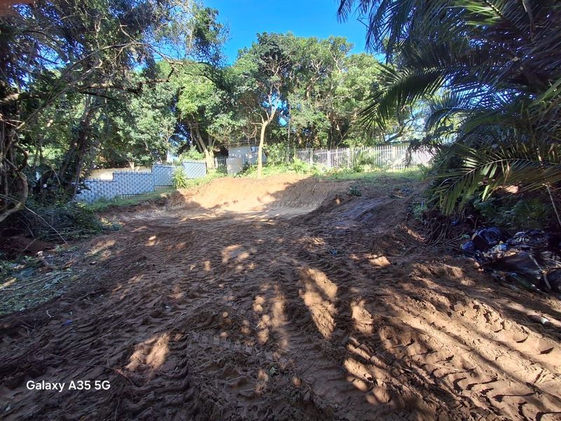 A view of a vacant residential plot featuring cleared earth, mature trees, and perimeter fencing, suitable for future development.