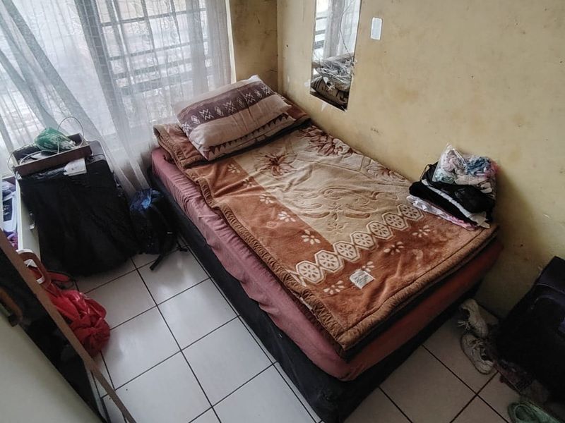 A compact bedroom featuring tiled flooring, a window with security bars, and a recessed wall mirror, currently requiring cosmetic wall repairs and repainting.