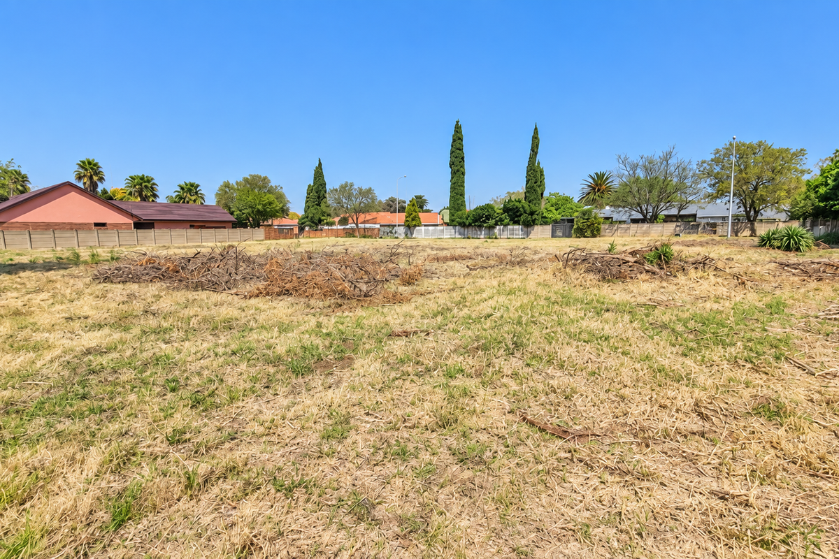 A vacant residential plot featuring dry grass, perimeter walling, and surrounding mature trees in a established neighborhood setting.