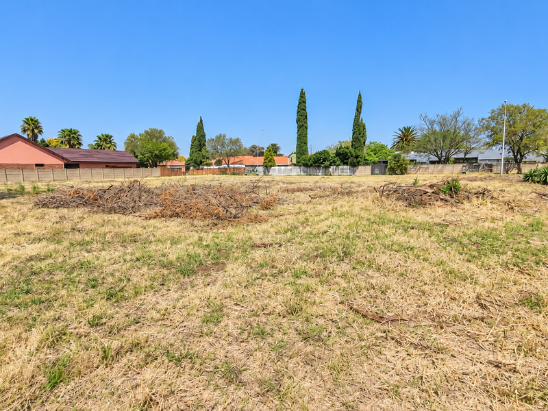 A vacant residential plot featuring dry grass, perimeter walling, and surrounding mature trees in a established neighborhood setting.