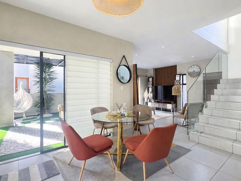 Bright open-plan living and dining area featuring tiled flooring, a modern glass-railed staircase, and large sliding doors providing seamless access to the patio.