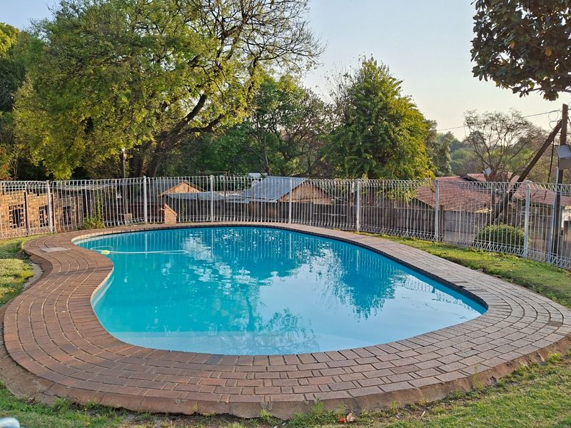 A well-maintained kidney-shaped swimming pool featuring brick coping and a secure metal perimeter fence, set within a landscaped garden area.