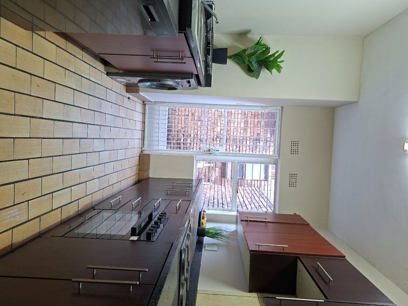 A functional kitchen featuring dark wood cabinetry, built-in oven and hob, tiled splashback, and a window with security bars.