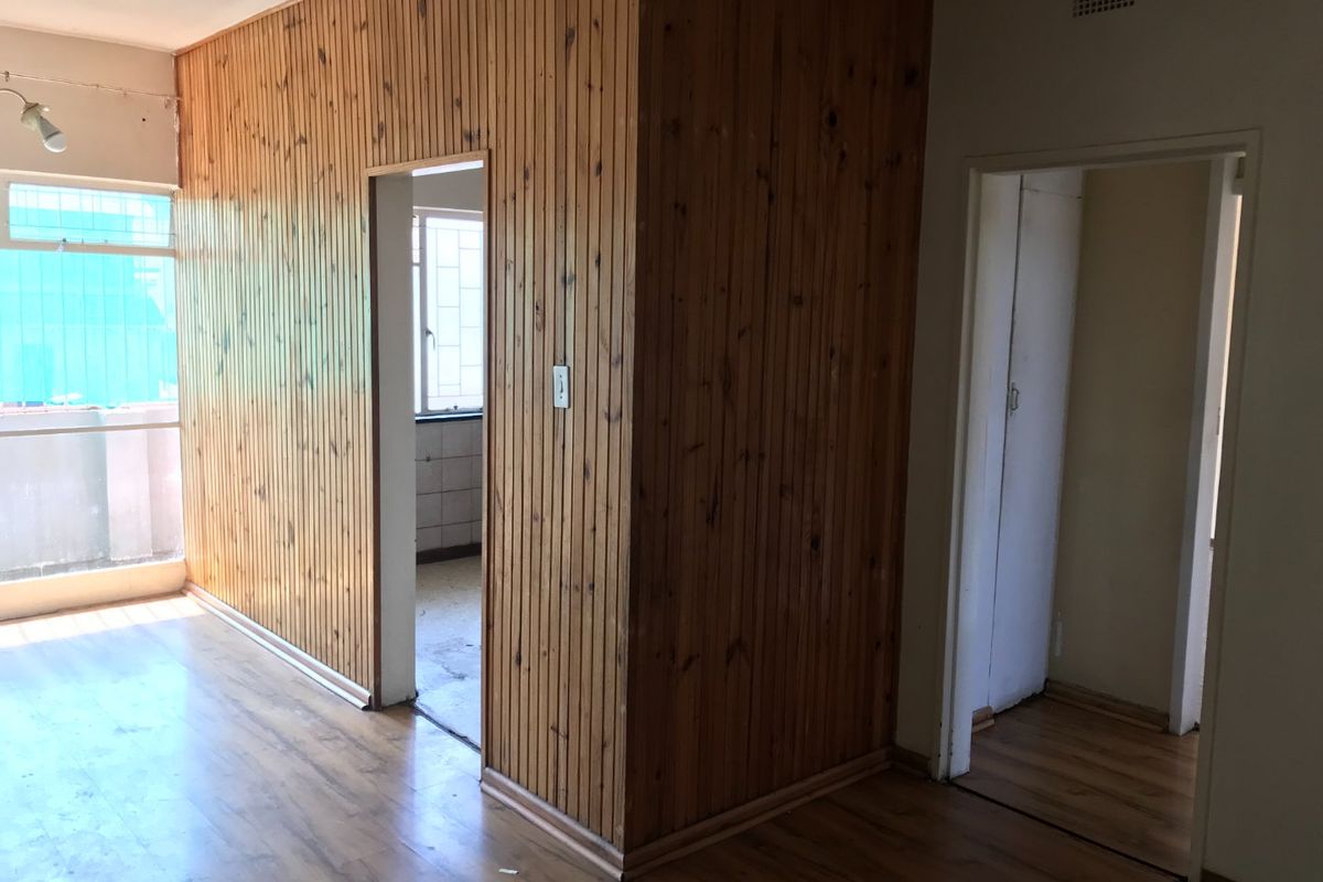 An empty living area featuring dated timber wall paneling and laminate flooring, with access to adjacent rooms and a balcony.