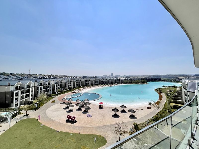 Elevated view overlooking a large communal swimming lagoon, sandy beach area, and surrounding apartment complex architecture.