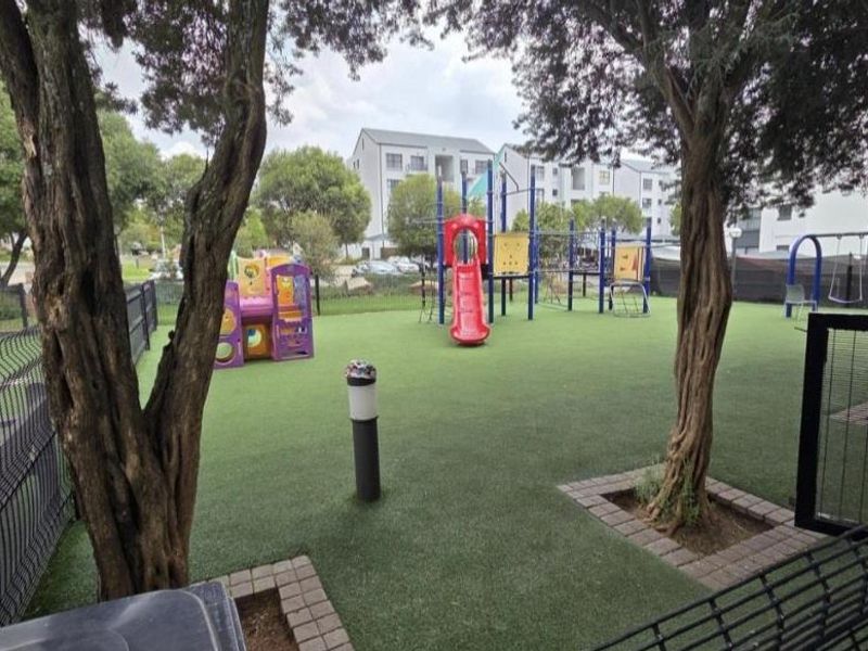 A well-maintained communal playground area featuring artificial turf, modern play equipment, and secure perimeter fencing within the apartment complex grounds.