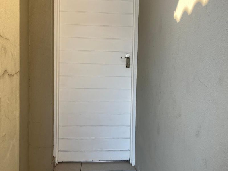 A narrow exterior entrance area featuring a white door and tiled flooring, showing visible cracks in the surrounding wall plaster.