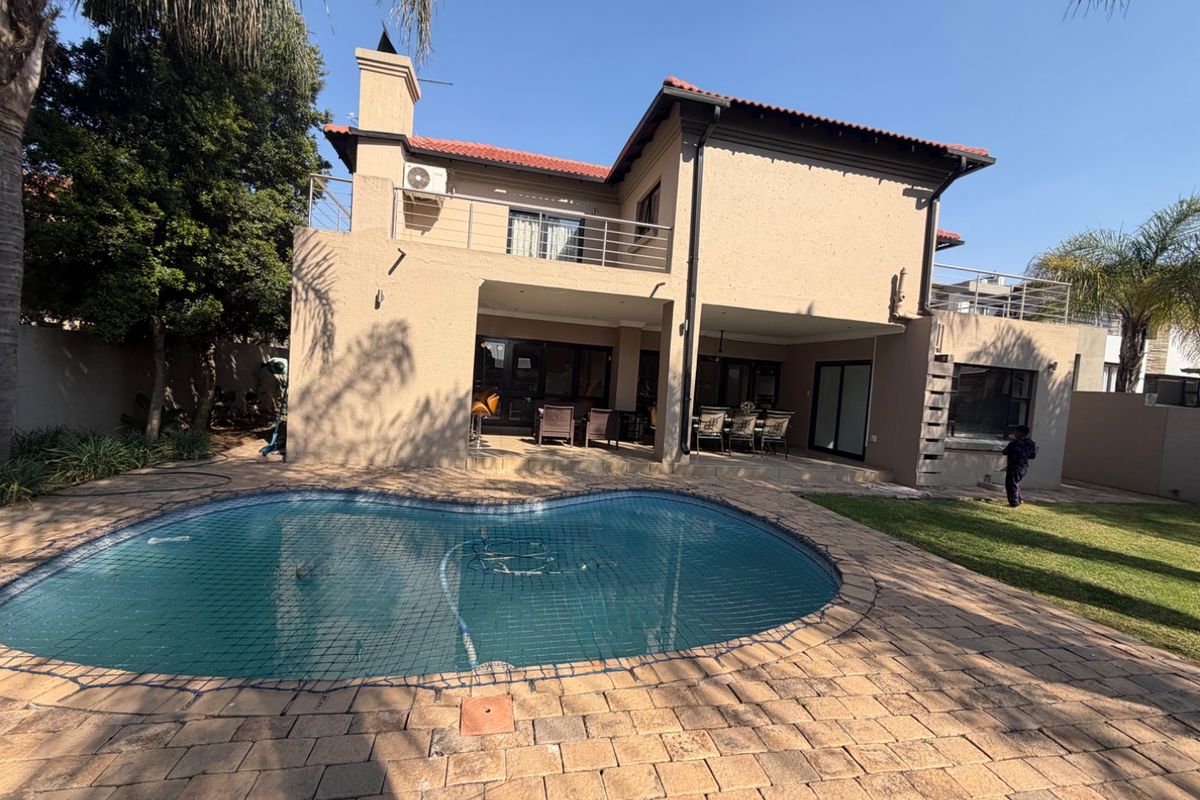 Rear view of a two-story house featuring a swimming pool, covered patio, upper-level balcony, and paved outdoor entertainment area.