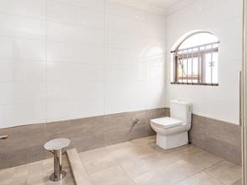 A clean, modern bathroom featuring neutral-toned wall and floor tiles, a white toilet, and an arched window with security bars.