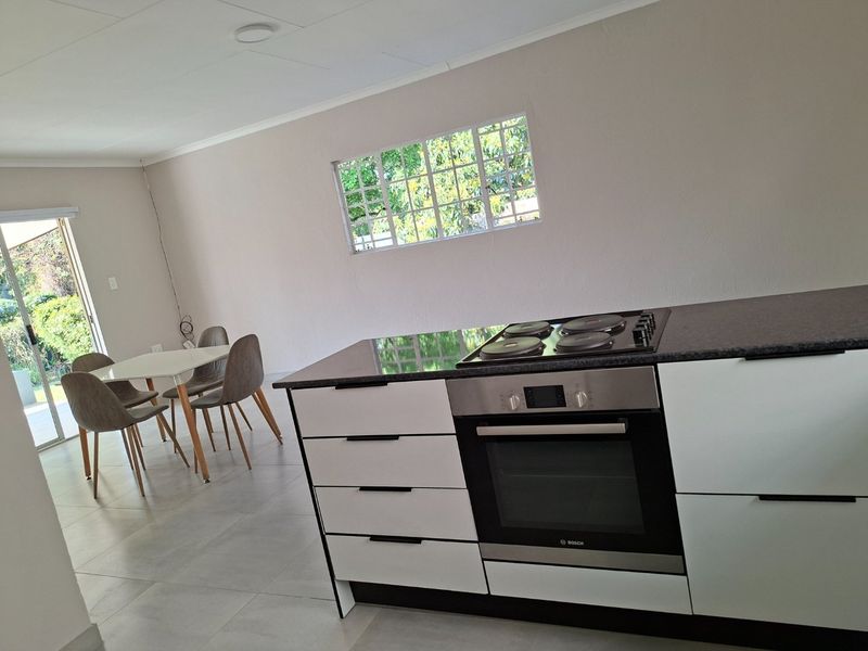Modern open-plan kitchen featuring sleek white cabinetry, dark countertops, built-in oven and hob, flowing into a bright dining area with garden access.