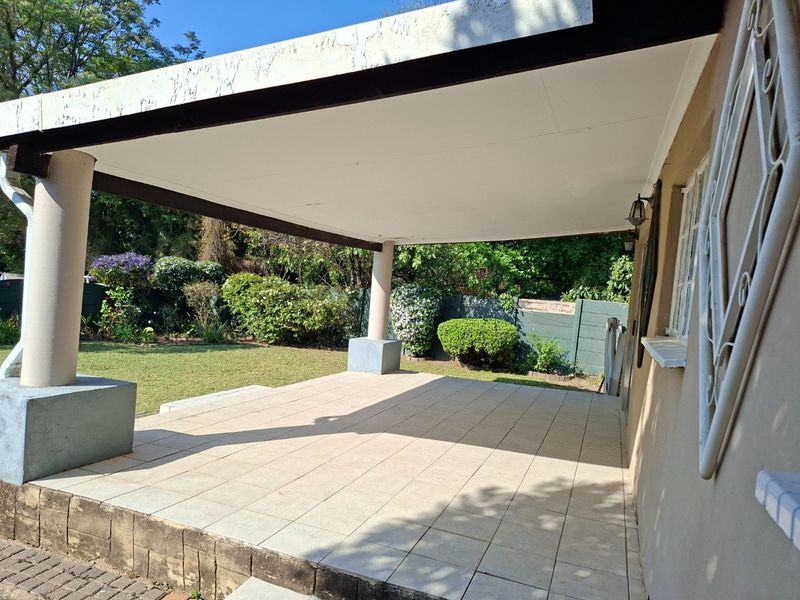 A spacious covered patio featuring tiled flooring and structural support columns, overlooking a private garden area.