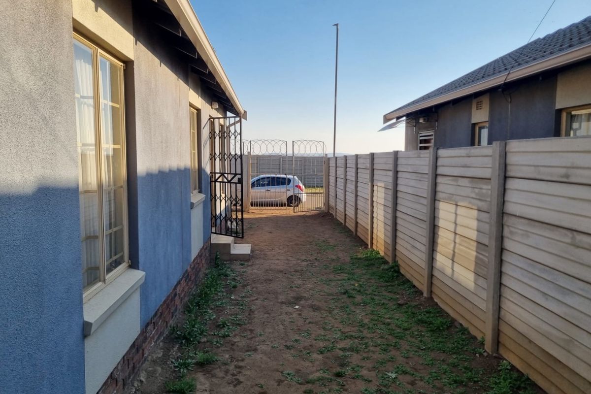 A side passage of the property featuring a precast concrete boundary wall, security gate with razor wire, and unlandscaped ground area.