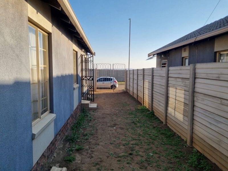 A side passage of the property featuring a precast concrete boundary wall, security gate with razor wire, and unlandscaped ground area.