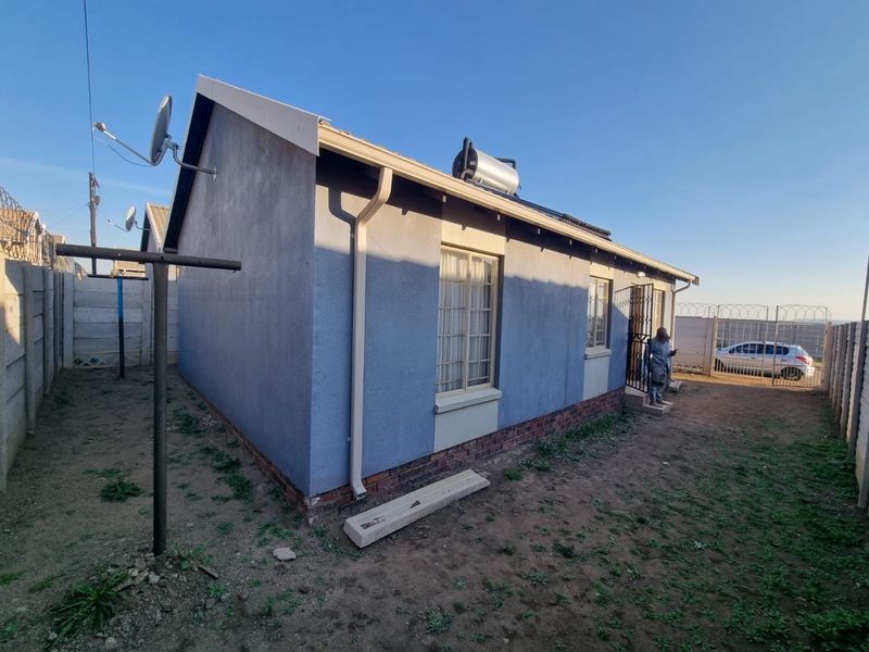 Rear view of the house featuring a solar water heater, rain gutters, and a secure fenced perimeter with a brick plinth foundation.