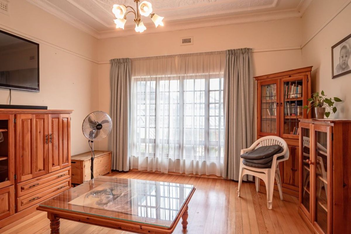 A living room featuring polished timber flooring, decorative ceiling detailing, and a large window with curtain tracks, offering a classic and well-maintained space.