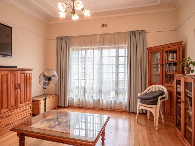A living room featuring polished timber flooring, decorative ceiling detailing, and a large window with curtain tracks, offering a classic and well-maintained space.