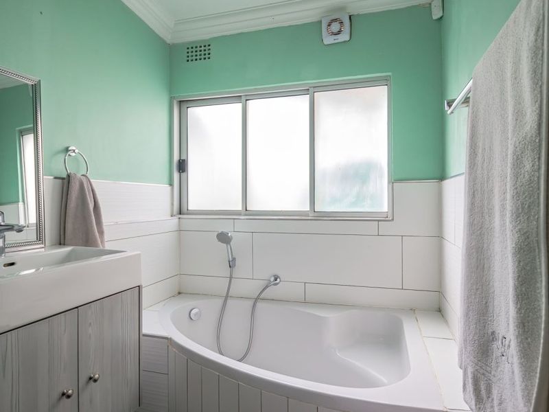 A functional bathroom featuring a corner bathtub with handheld shower, a modern vanity with basin, and tiled splashbacks in a light green color scheme.
