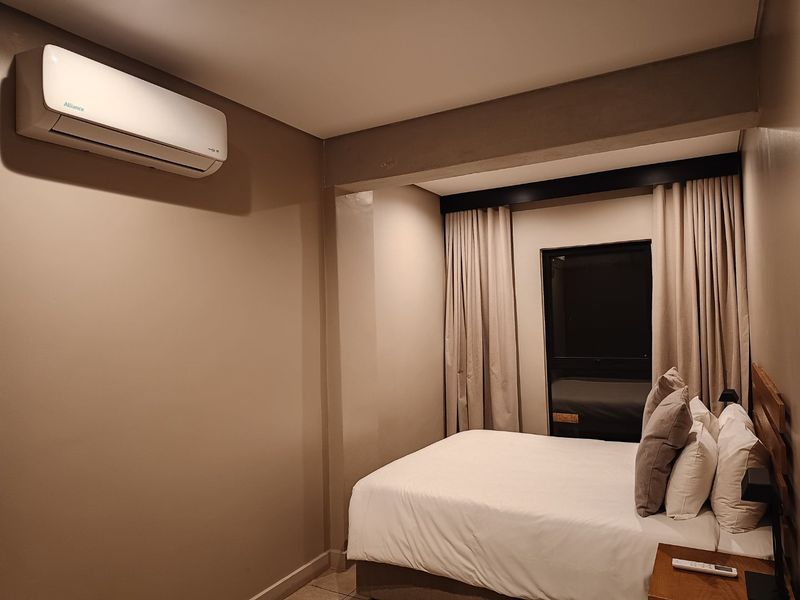 A well-maintained bedroom featuring a wall-mounted air conditioning unit, neutral wall tones, and floor-to-ceiling curtains.