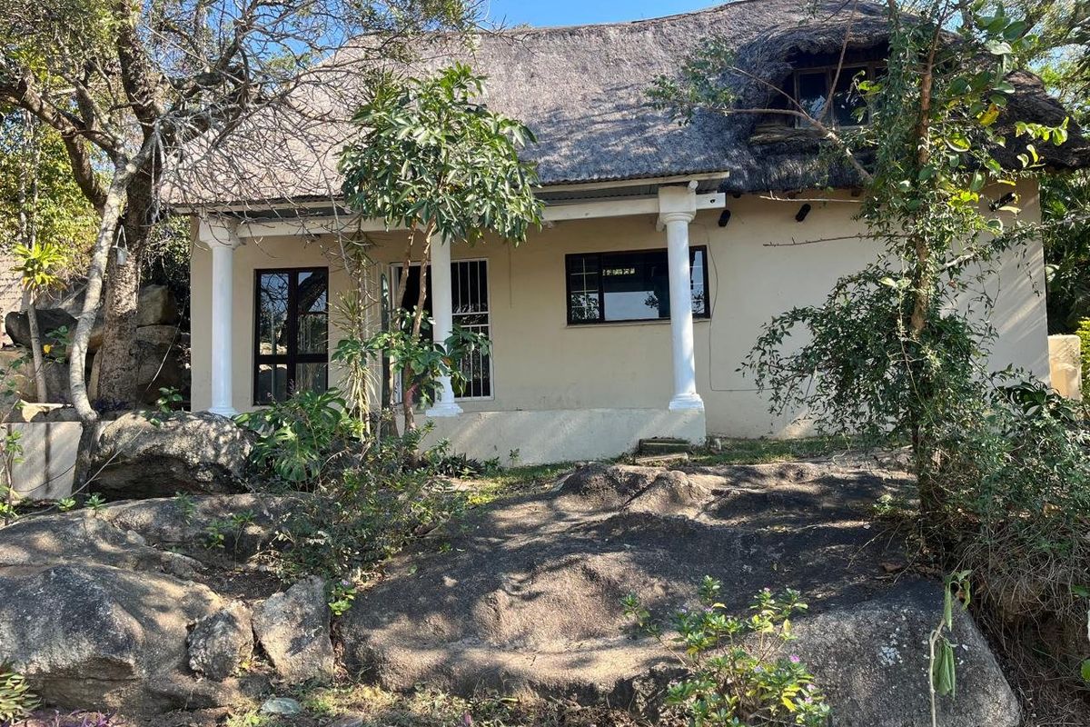 Rear view of a rustic farm-style cottage featuring a traditional thatched roof, covered patio with classical columns, and natural rocky landscape surroundings.
