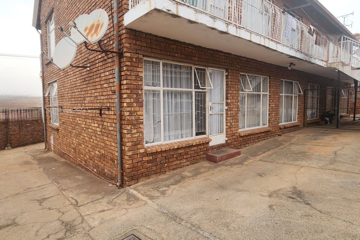 Exterior view of a multi-story apartment building featuring face brick construction, a concrete balcony with metal railings, and paved ground level access.